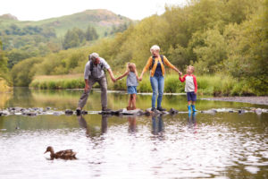 Grandparents,With,Grandchildren,Crossing,River,Whilst,Hiking,In,Uk,Lake